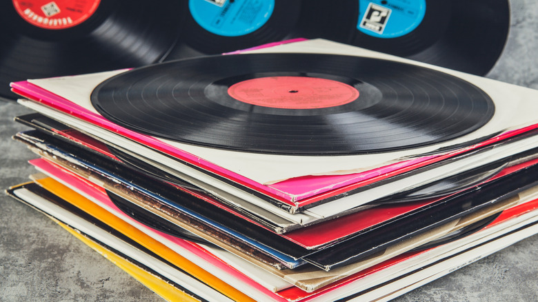 A colorful stack of vinyl records, the records are laying on top of grey carpet, and one single record sits on top of the stack to hide the cover of the one under it