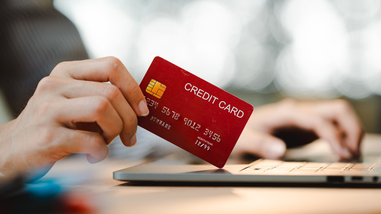 A closeup of a red credit card held in a user's right hand as they type on a laptop with their left, the face of the individual cannot be seen