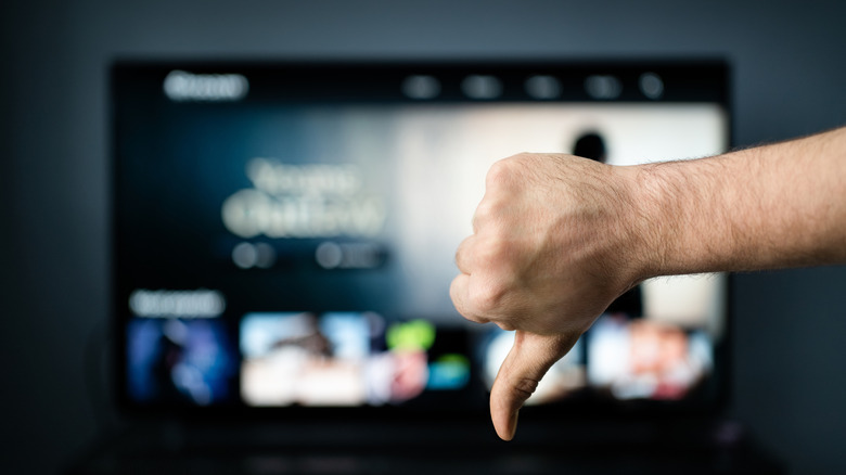 Person giving a thumbs-down gesture in front of a blurred streaming menu on a TV.