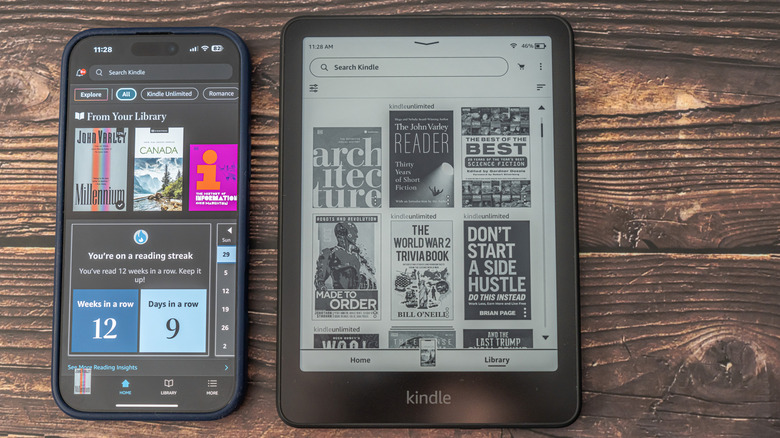 A Kindle displaying its library alongside a smartphone running the Kindle app on a wooden surface
