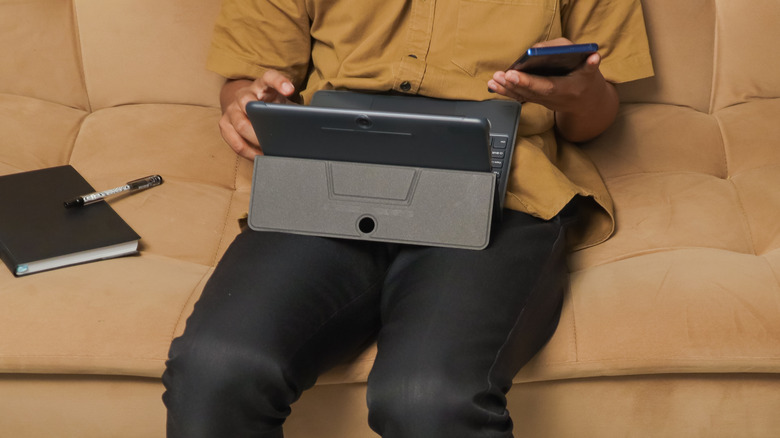 Person sitting on a couch using a tablet with a keyboard case while holding a smartphone.