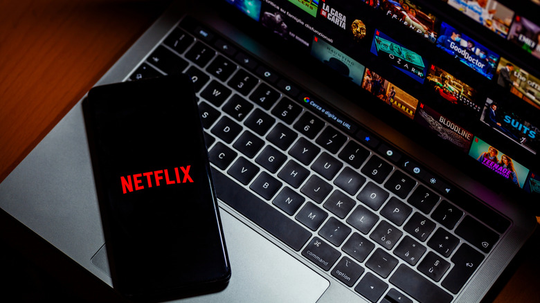 Netflix logo displayed on a smartphone beside a laptop showing streaming titles.