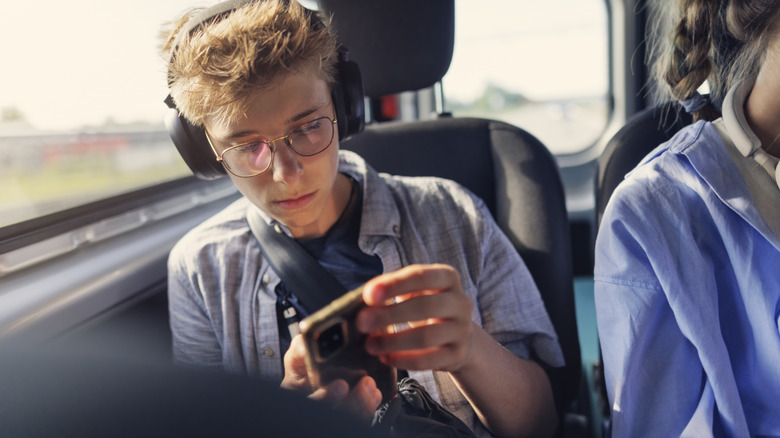 A person in a blue unbuttoned shirt wears black headphones and has both their hands around a smartphone as they sit on what appears to be a bus