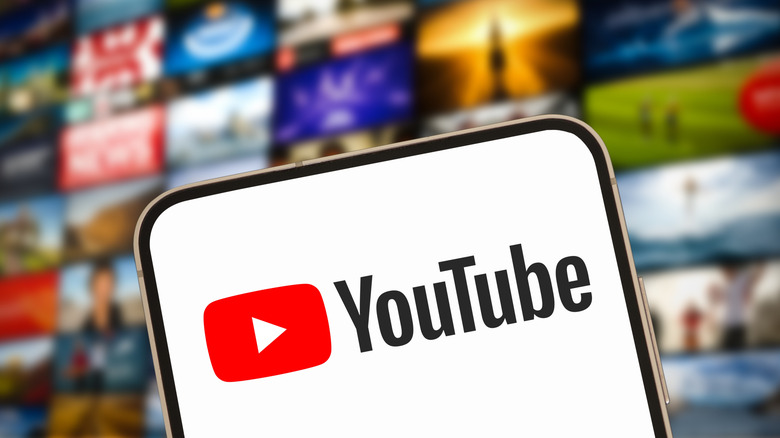 A smartphone displaying the YouTube logo sits in front of a wall of heavily blurred images, suggesting they are thumbnails for YouTube videos