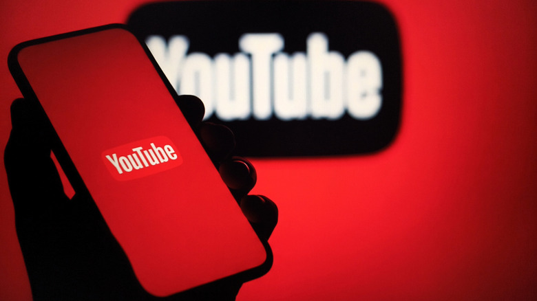 A smartphone displaying the YouTube logo against a red background is held up in front of a red backdrop that is also showing a blurred YouTube logo in black