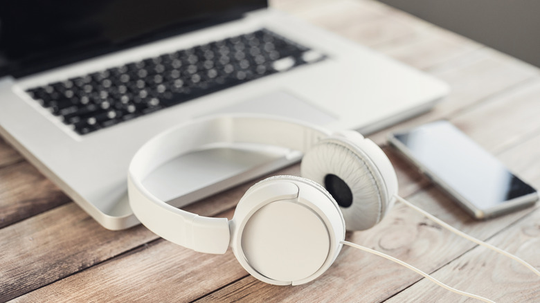 A pair of white wired headphones sits on the edge of a silver laptop next to a smartphone, all three items are on a wooden surface