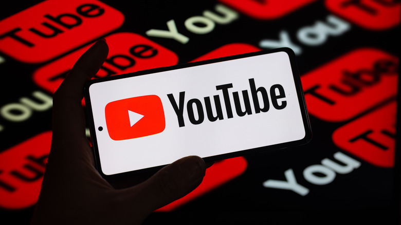 A heavily shadowed hand holds a smartphone displaying the YouTube logo against a patterned background of darker YouTube logos