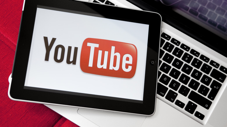 A tablet displaying the YouTube logo sits on top of a silver laptop, both items are on a red fabric surface