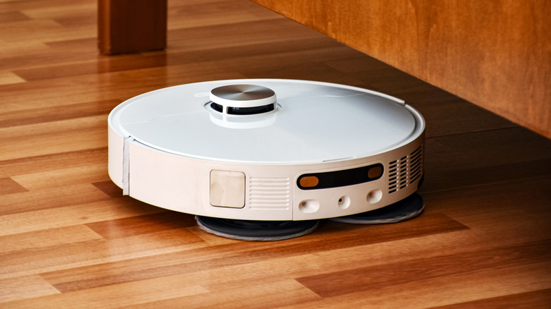 White robot vacuum cleaning a wooden floor near furniture.