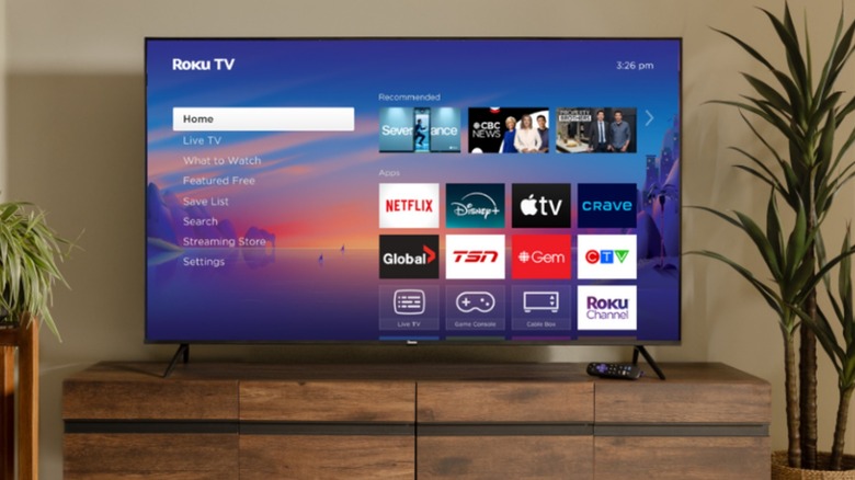 A Roku TV displaying its home screen with a multitude of streaming apps sits on top of a wooden TV stand, there are plants on each side of the display