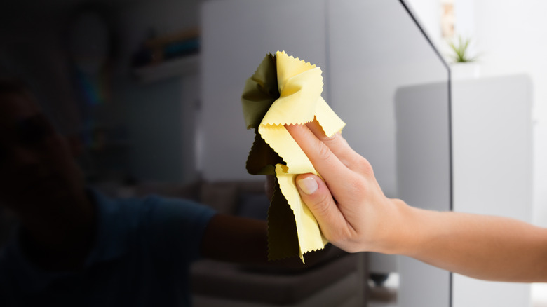 A human hand uses a microfiber cloth to wipe down a TV display, while only a portion of the room can be seen, the room appears to be mostly white in color