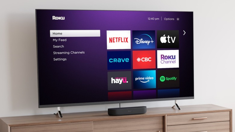 An older model Roku TV displaying its Home screen with several streaming apps sits on top of a brown TV stand, at least one drawer from the stand can be seen
