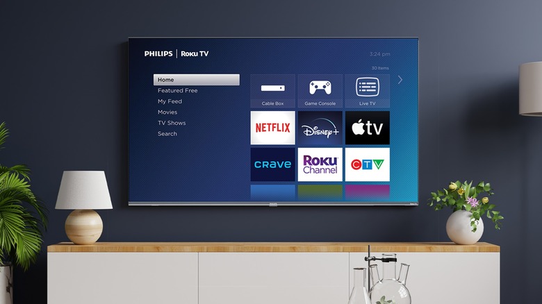 A Roku TV displaying its Home screen with several streaming apps is attached to a blue wall, there is a white TV stand with a lamp and a potted plant sitting on top of it