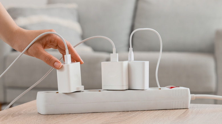 A white surge protector sits on the edge of a table in front of a grey couch, one hand can be seen placing a charging block into one of the protector's ports