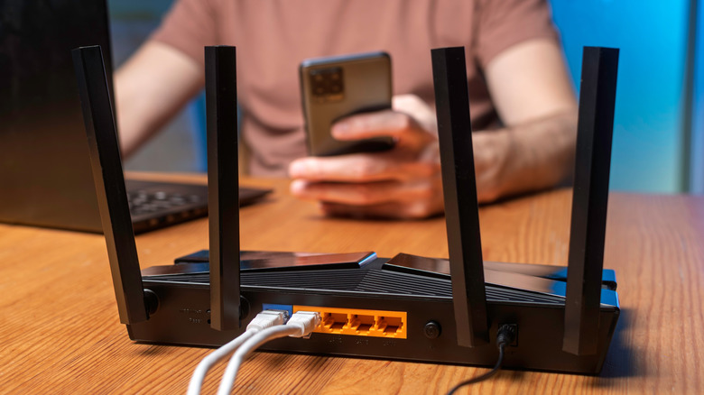 A router on a table with a man in the background on his phone and laptop as if he is configuring it
