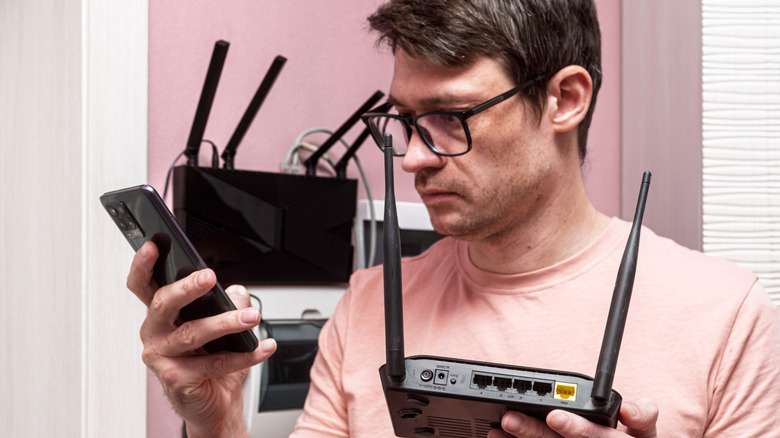 A man holding a router in one hand while looking at a phone he is holding in the other hand