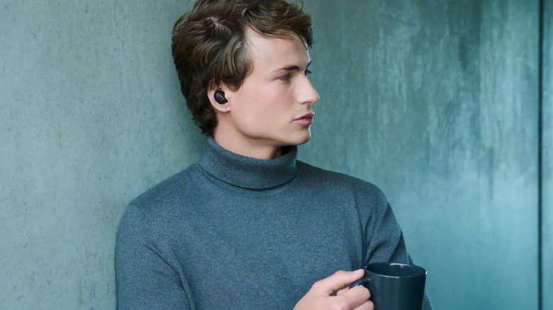 Person wearing Sony WF-1000XM6 noise-canceling earbuds while leaning against wall and holding cup