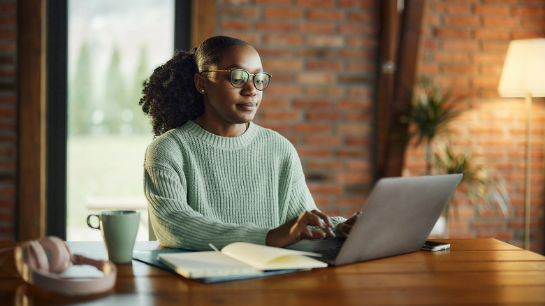 A woman sitting at a table typing on a laptop with headphones, a mug, and an open notebook in front of her