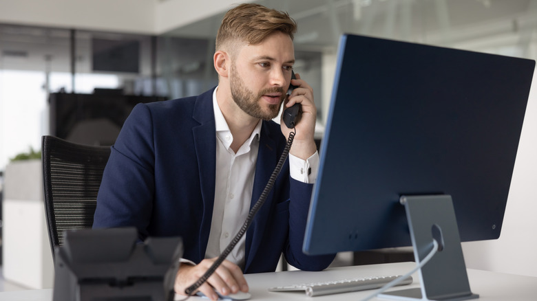 A man in a suit sitting at a desk talking into a phone and looking at a computer monitor