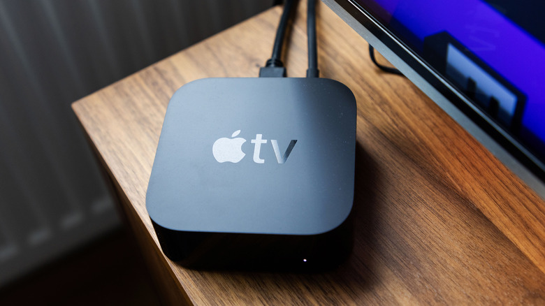 A black Apple TV streaming device sits near the corner of a brown, wooden TV stand
