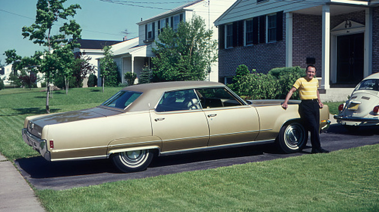 A man leans against a 1960s vehicle in front of his house