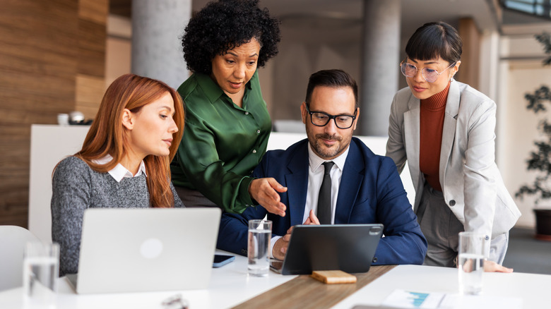 A group of individuals dressed in business attire gather around a laptop screen, two people are sitting and two are standing