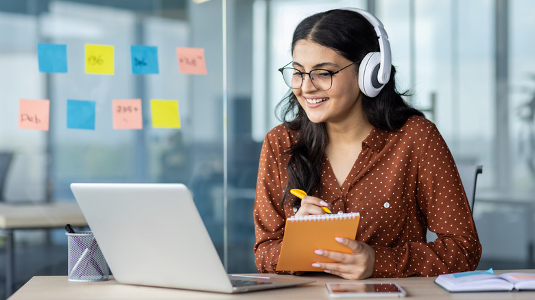 A woman wearing white headphones and glasses sits as a desk in front of a laptop, she is holding a pen and notebook in her hands, seemingly to take notes