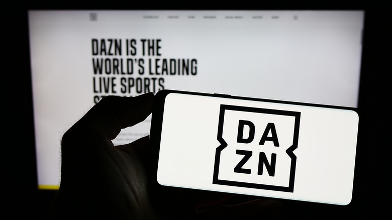 Person holding mobile phone with logo of DAZN on screen in front of web page