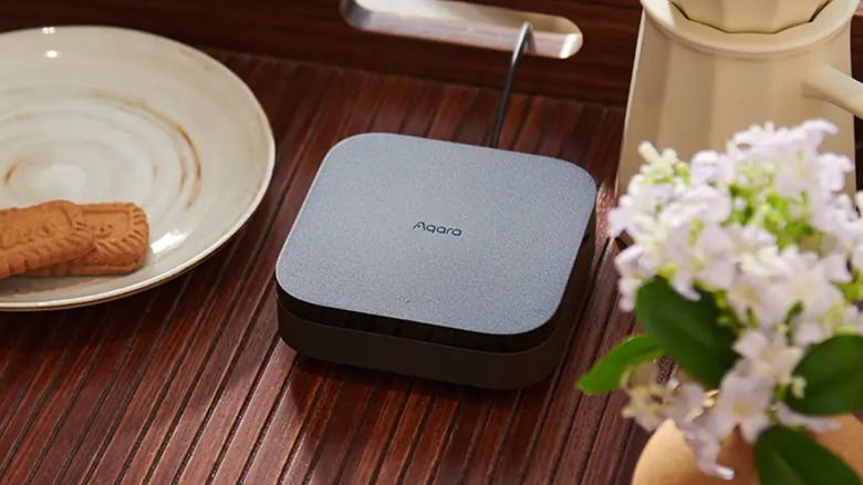 Aqara Hub M3 on a wooden table beside a plate and flowers.