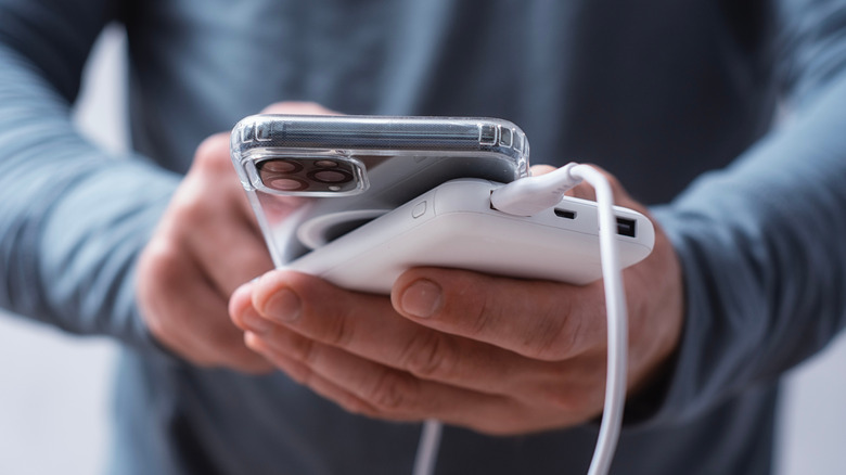 A closeup of an individuals hands as they hold a smartphone connected via USB to a white charging power bank