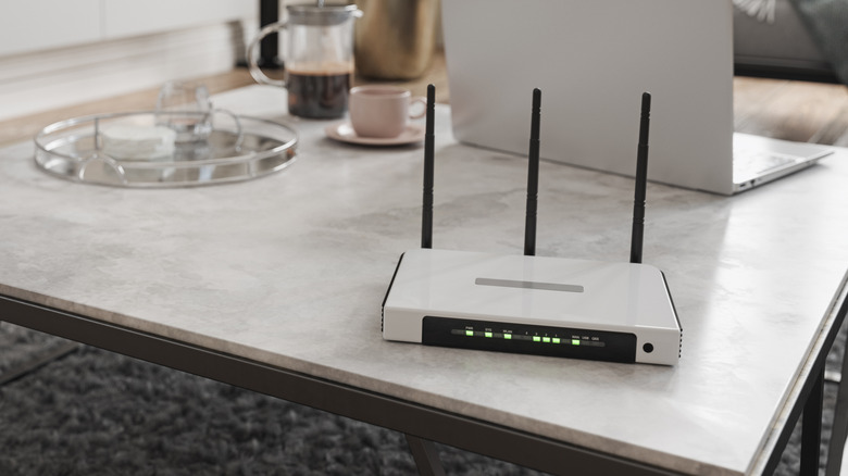 A white router sits on a table that has a white marble surface, a silver laptop sits behind the router alongside a cup of coffee and a glass serving tray
