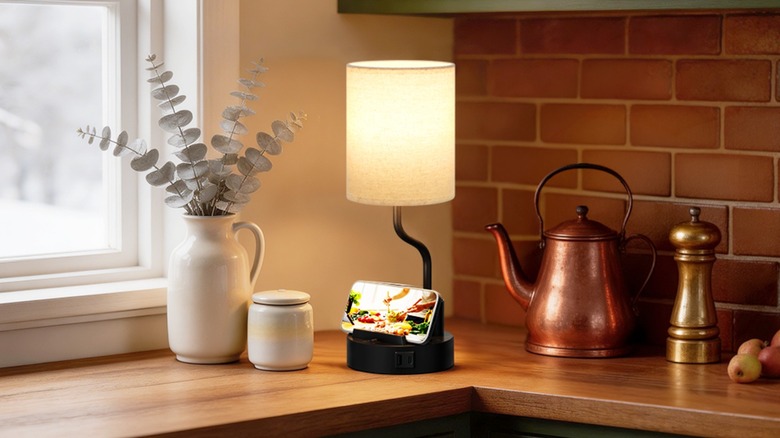 The Fenmzee Bedside Table Lamp with USB Charging sitting on a kitchen counter, a smartphone is nestled in the lamps' smartphone holding slot