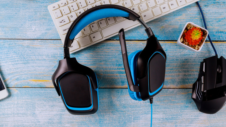 A black gaming headset with microphone sits on a blue wooden surface next to a black gaming mouse, a white keyboard, and a small potted plant