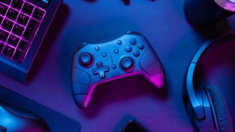 A black game controller with two joysticks and a D-pad sits on a desk between an RGB keyboard and a pair of black headphones
