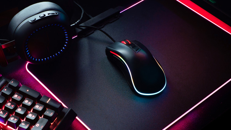 A black gaming mouse with a red glow sits on a black mousepad next to a pair of black headphones and keyboard
