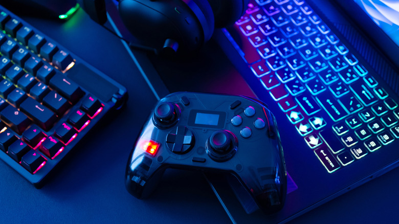 A gaming controller with two different joysticks and a D-pad sits in between a laptop with a RGB keyboard and another RGB keyboard