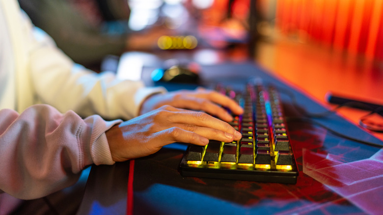 A user with two hands on an RGB keyboard that's glowing yellow in the photo, the keyboard sits on a cushion that is on a desk