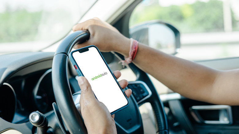 Android Auto in print displayed on a phone while someone is holding on to a steering wheel inside a car