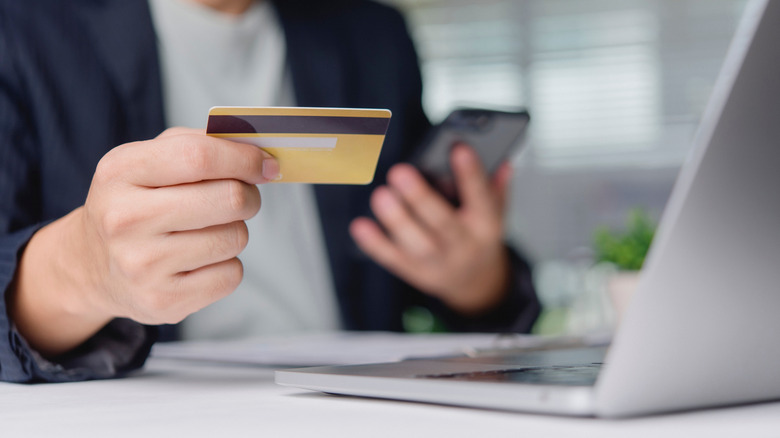 person holding a credit card in one hand, smartphone in another, with a laptop placed in the front