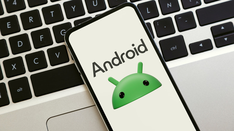 The Android OS icon symbolized on a smart phone screen, set on top of a laptop keyboard.