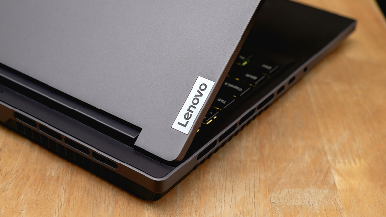 A close-up of a Lenovo laptop partially open on a wooden table, showing the logo and side vents.