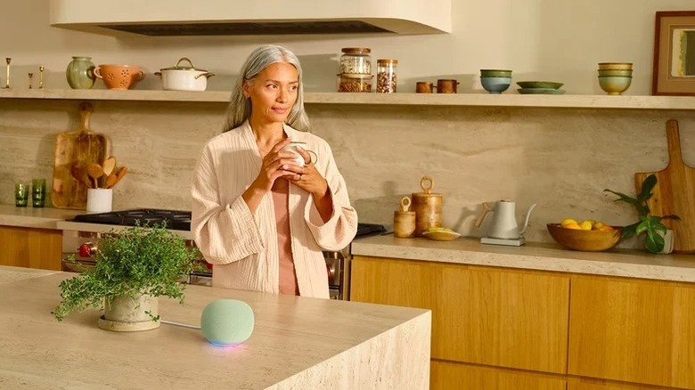 A woman sipping coffee in a kitchen with a Google Home speaker on the counter.