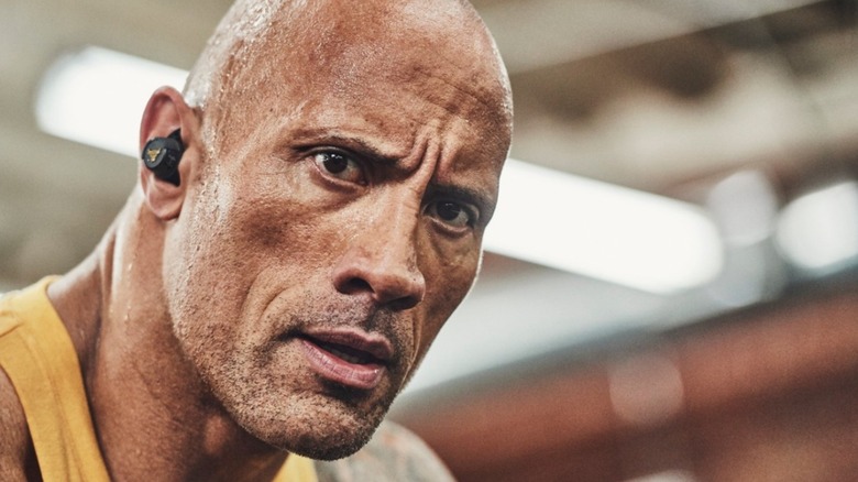 Dwayne "The Rock" Johnson wearing a pair of black in-ear Project Rock Training headphones as he looks directly at the camera with a stern expression
