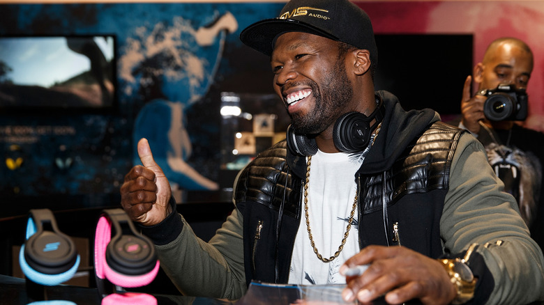 Rapper 50 Cent sits next to two pairs of SMS Audio headphones. the artist is also notably wearing an SMS Audio hat that is black and gold