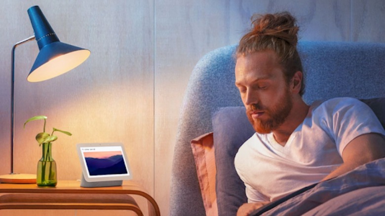 google Nest Hub on a bedside table under a warm lit lamp with a sleepy man giving a voice command.