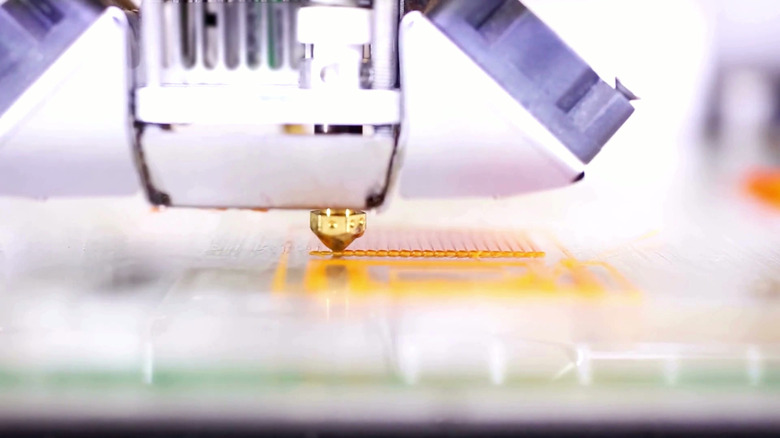 3D printer nozzle laying orange filament on a print bed.
