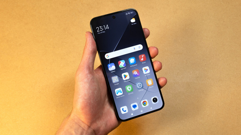 The left hand of a person holding a Xiaomi smartphone, the phone is displaying its Home screen, which shows 12 different apps