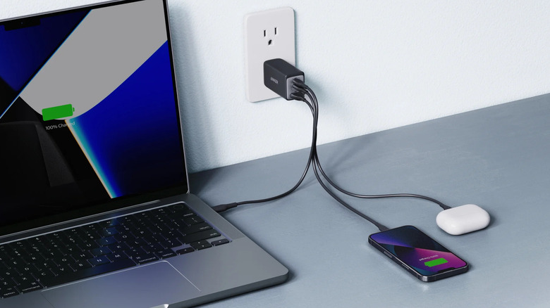 Anker 735 wall charger plugged into an outlet, charging a laptop, smartphone, and wireless earbuds on a desk.