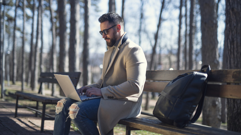 A person wearing sunglasses sitting on a park bench and working on a laptop