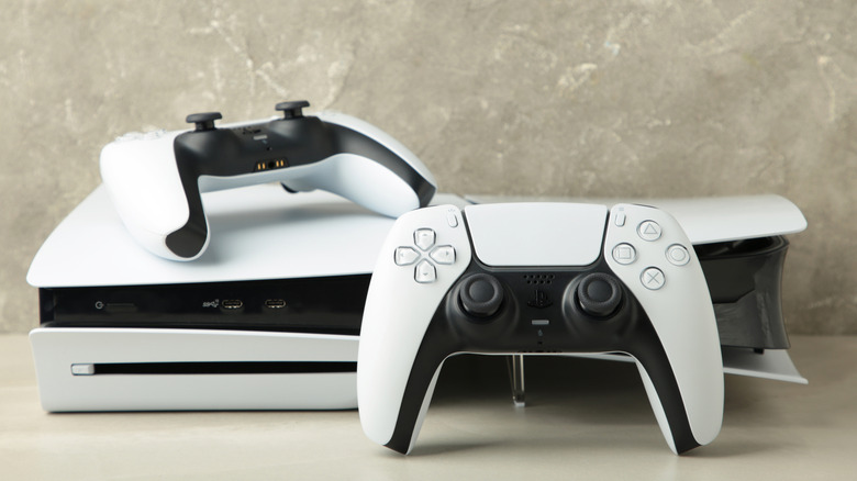 A white PlayStation 5 with two Sony Dual Sense wireless controllers, one is sitting in front of the console and the other sits on top of the console